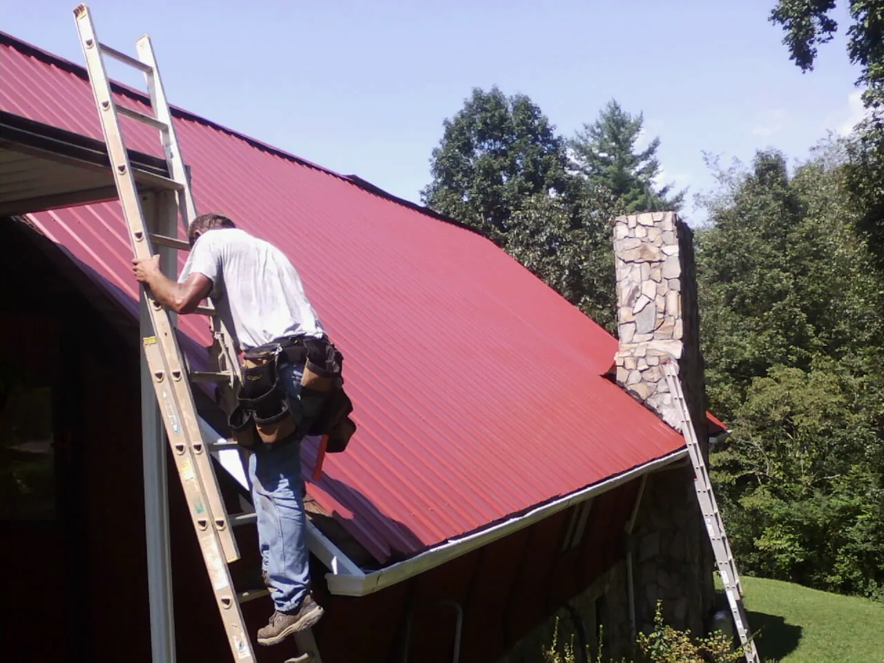 Roofer on a ladder beside a red metal roof and stone chimney — Soffit & Fascia Repair by Roofing Brothers in Rudolph